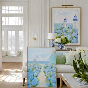 May include: Two framed paintings with coastal scenes, featuring blue hydrangeas and lighthouses. The artwork is displayed in a bright, white-walled room with a white sofa, a coffee table, and decorative vases. The paintings have gold frames.
