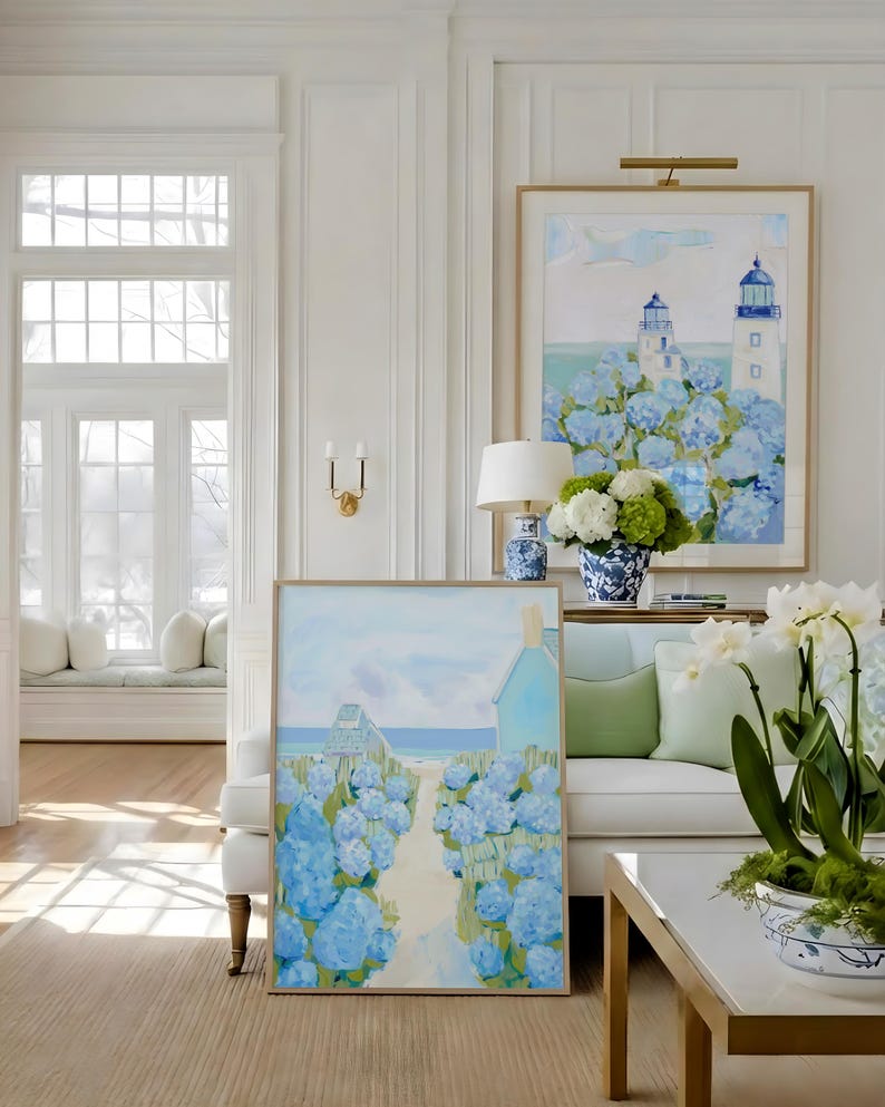 May include: Two framed paintings with coastal scenes, featuring blue hydrangeas and lighthouses. The artwork is displayed in a bright, white-walled room with a white sofa, a coffee table, and decorative vases. The paintings have gold frames.