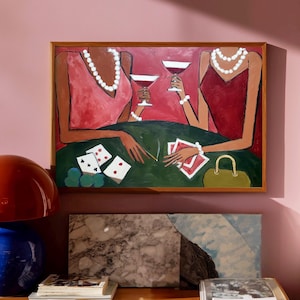 May include: An abstract painting depicts two figures in dresses, playing cards and drinking cocktails. The figures are adorned with pearl necklaces and bracelets. The painting is framed in a wooden frame and hangs on a pink wall. A green table is in the foreground.