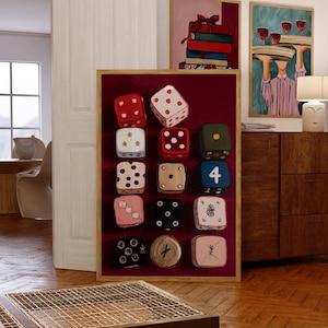 Op de afbeelding: Een ingelijst kunstwerk met twaalf dobbelstenen met verschillende puntconfiguraties, tegen een dieprode achtergrond. De dobbelstenen zijn in rood, wit, zwart en beige. Twee andere ingelijste schilderijen zijn zichtbaar op de achtergrond.