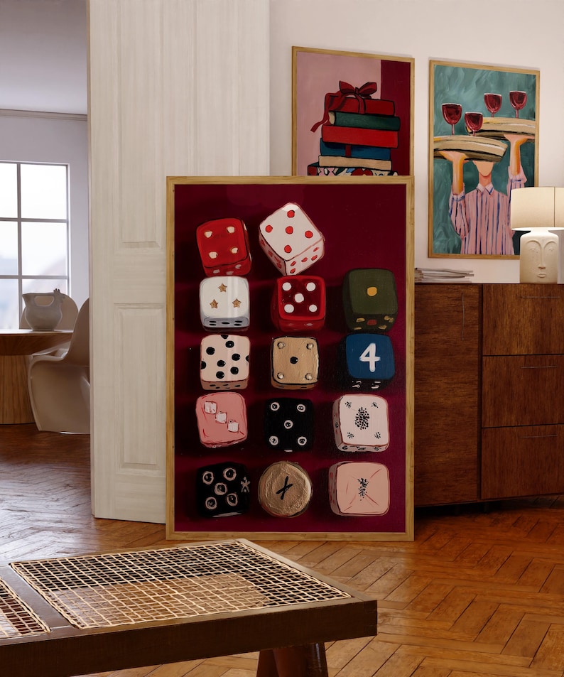 Op de afbeelding: Een ingelijst kunstwerk met twaalf dobbelstenen met verschillende puntconfiguraties, tegen een dieprode achtergrond. De dobbelstenen zijn in rood, wit, zwart en beige. Twee andere ingelijste schilderijen zijn zichtbaar op de achtergrond.