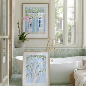 Puede incluir: Dos pinturas de acuarela enmarcadas en un ba&ntilde;o. Una representa un edificio con contraventanas azules, la otra un &aacute;rbol con flores azules. El ba&ntilde;o tiene una ba&ntilde;era blanca, baldosas verdes y una ventana.