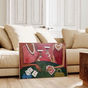 May include: An art print depicting two figures in red dresses, adorned with pearls and bracelets, holding wine glasses. The scene includes a green table with playing cards and poker chips. A gold frame surrounds the artwork, which is leaning against a beige sofa.