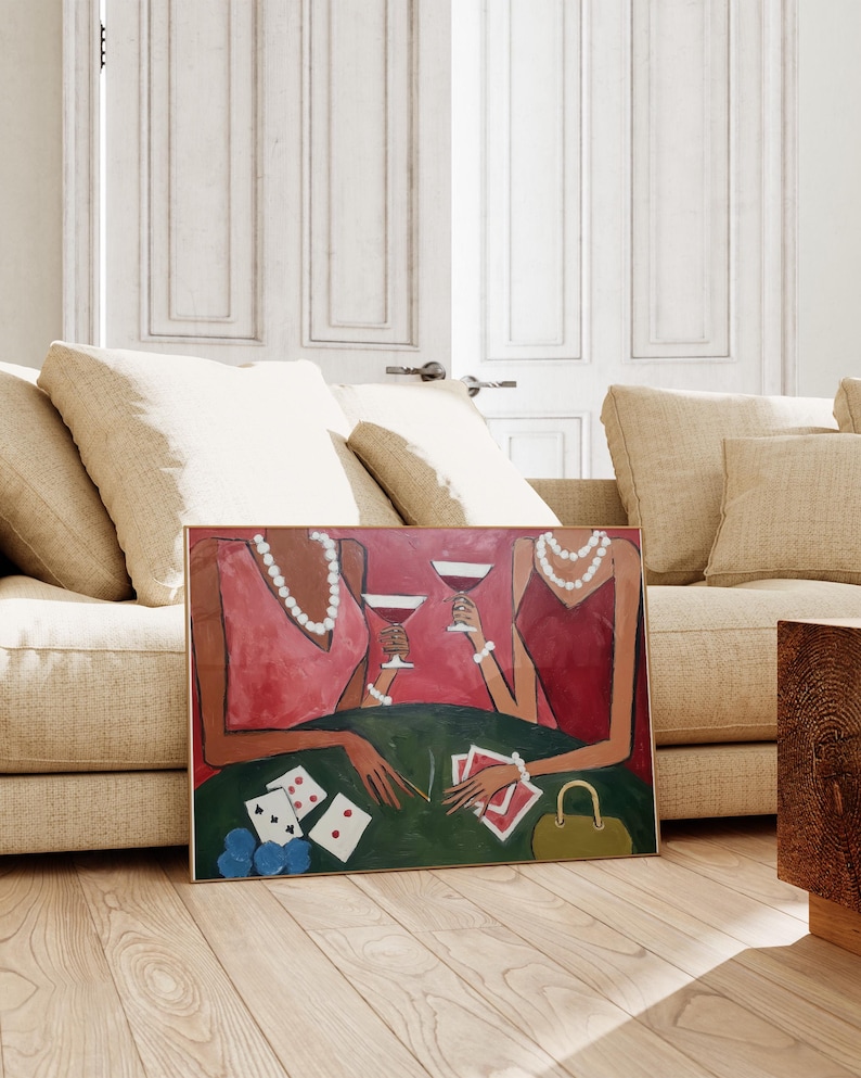 May include: An art print depicting two figures in red dresses, adorned with pearls and bracelets, holding wine glasses. The scene includes a green table with playing cards and poker chips. A gold frame surrounds the artwork, which is leaning against a beige sofa.