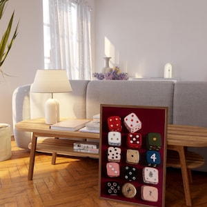 Op de afbeelding: Een ingelijste display van twaalf decoratieve dobbelstenen met verschillende kleuren en patronen. De dobbelstenen staan tegen een bordeauxrode achtergrond in een houten frame. De dobbelstenen hebben verschillende nummers en ontwerpen, waaronder stippen, sterren en andere symbolen.