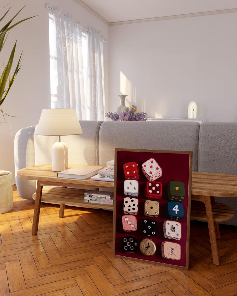 Op de afbeelding: Een ingelijste display van twaalf decoratieve dobbelstenen met verschillende kleuren en patronen. De dobbelstenen staan tegen een bordeauxrode achtergrond in een houten frame. De dobbelstenen hebben verschillende nummers en ontwerpen, waaronder stippen, sterren en andere symbolen.