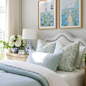 May include: A bedroom scene featuring a bed with light blue bedding and decorative pillows. Two framed watercolor paintings of lighthouses and blue hydrangeas hang above the bed. A white lamp and a blue and white vase are on a nightstand.