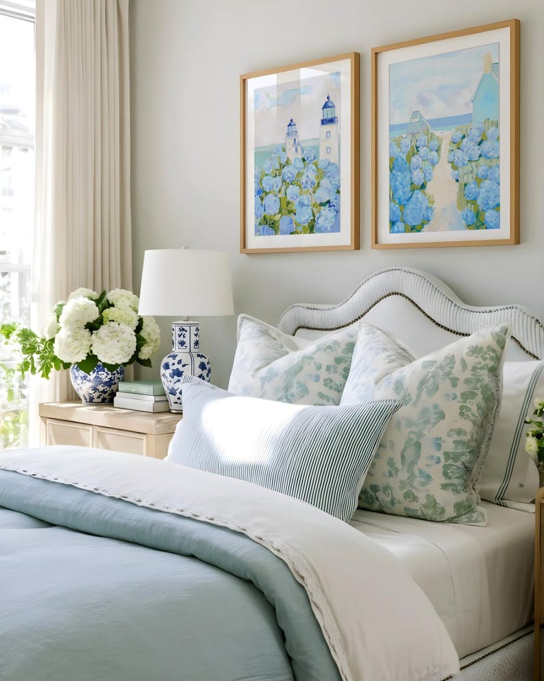 May include: A bedroom scene featuring a bed with light blue bedding and decorative pillows. Two framed watercolor paintings of lighthouses and blue hydrangeas hang above the bed. A white lamp and a blue and white vase are on a nightstand.