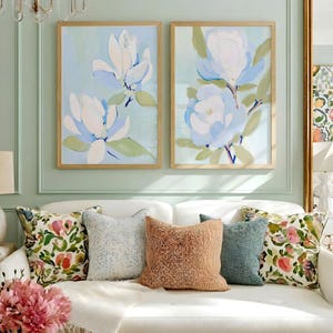 May include: Two framed floral art prints with light blue backgrounds and white and blue flowers hang above a white sofa with decorative pillows. The pillows include floral and textured designs in various colors.