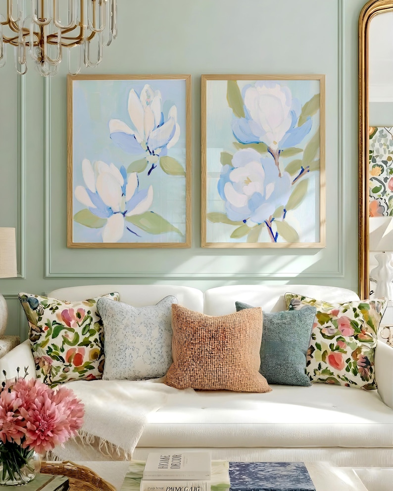 May include: Two framed floral art prints with light blue backgrounds and white and blue flowers hang above a white sofa with decorative pillows. The pillows include floral and textured designs in various colors.