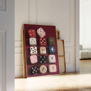Op de afbeelding: Een ingelijst kunstwerk met twaalf dobbelstenen met diverse ontwerpen en kleuren, tegen een dieprode achtergrond. De dobbelstenen bevatten stippen, sterren en andere symbolen. Het kunstwerk leunt tegen een muur.