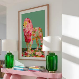 K&ouml;nnte beinhalten: Ein gerahmter Kunstdruck eines L&ouml;wen mit rosa und floraler M&auml;hne und K&ouml;rper, vor einem gr&uuml;nen Hintergrund. Das Kunstwerk wird &uuml;ber einem rosa Konsolentisch mit zwei gr&uuml;nen Glaslampen mit wei&szlig;en Schirmen ausgestellt. B&uuml;cher sind auf dem Tisch gestapelt.