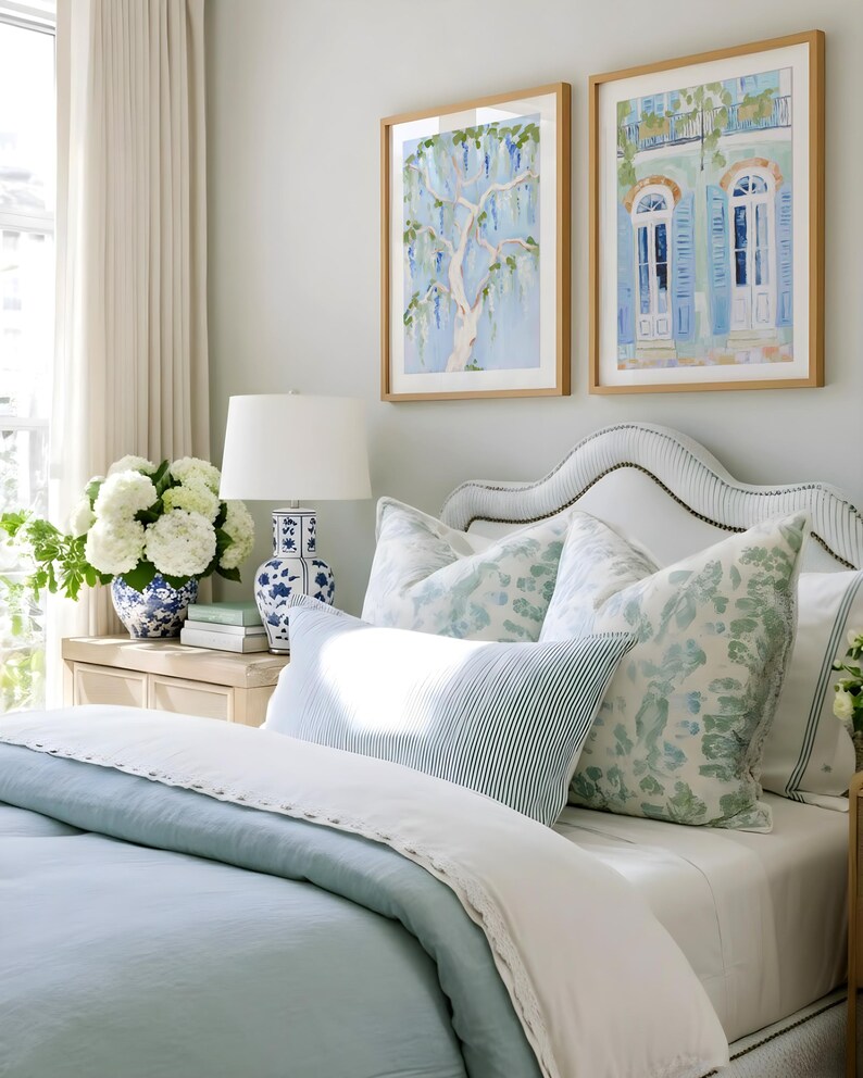 Puede incluir: Un dormitorio con una cama con ropa de cama azul claro y cojines estampados. Dos pinturas de acuarela enmarcadas cuelgan sobre la cama. Un jarr&oacute;n azul y blanco exhibe flores blancas.