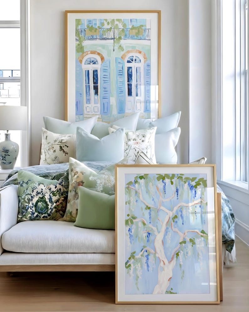 Puede incluir: Una escena interior luminosa con obras de arte enmarcadas y cojines decorativos. Las obras de arte incluyen una pintura de un &aacute;rbol con flores azules y una pintura de contraventanas azules. Los cojines son en tonos azules y verdes.