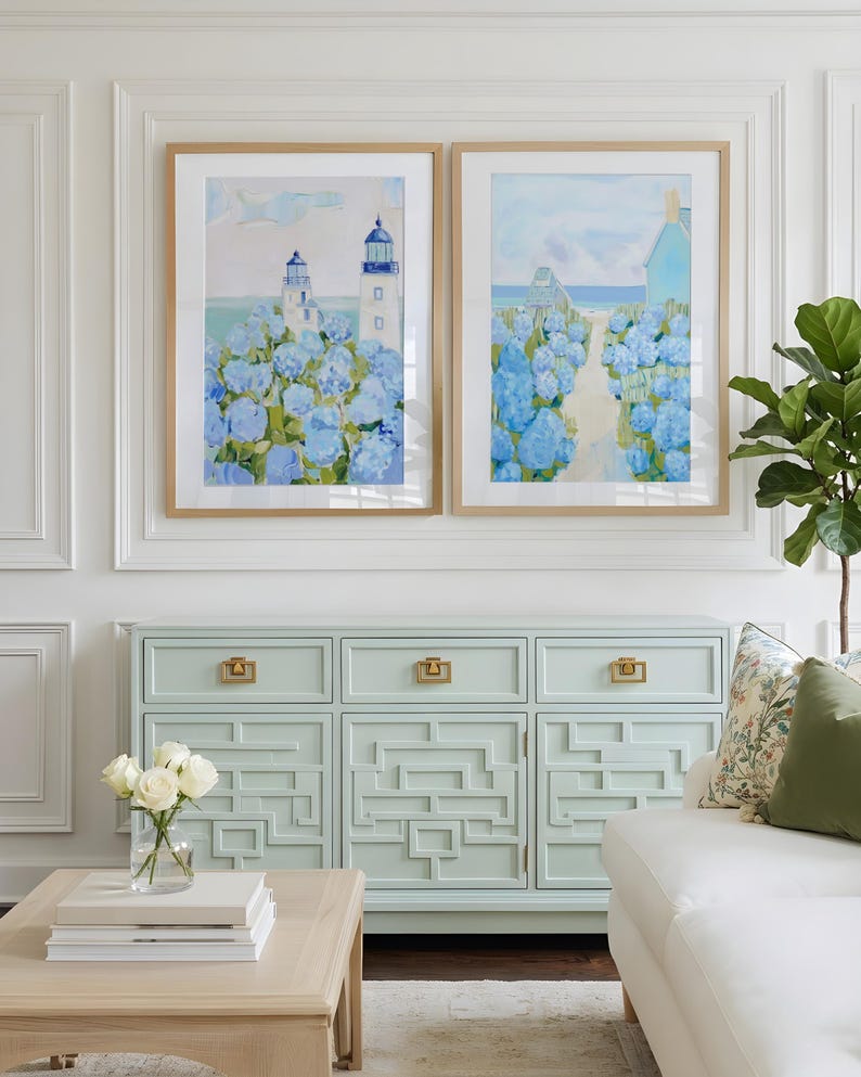 May include: Two framed watercolor paintings of coastal scenes with lighthouses and blue hydrangeas. Below, a light blue cabinet with gold hardware and a geometric design. A white sofa and a wooden coffee table complete the room.