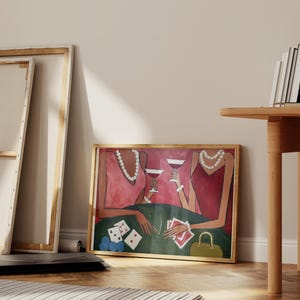 May include: A framed art print depicts two figures in red dresses at a poker table, holding wine glasses. The green table has playing cards and poker chips. A gold purse sits nearby. The artwork is in a wooden frame.