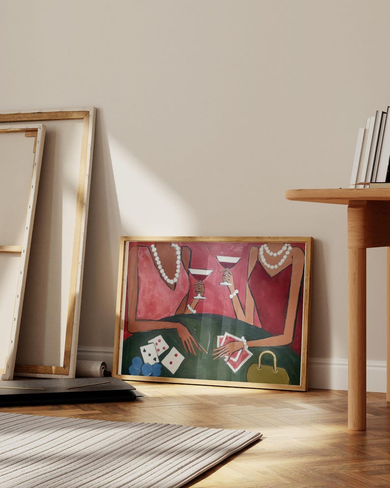 May include: A framed art print depicts two figures in red dresses at a poker table, holding wine glasses. The green table has playing cards and poker chips. A gold purse sits nearby. The artwork is in a wooden frame.