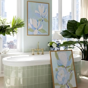 May include: A bathroom scene featuring a white bathtub with brass fixtures, a framed floral painting, and a large potted plant. The walls are a soft green, and the painting depicts white and blue flowers.