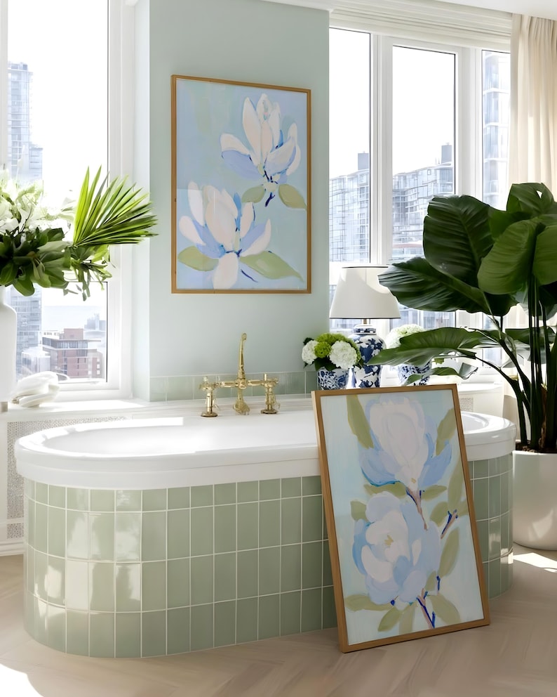 May include: A bathroom scene featuring a white bathtub with brass fixtures, a framed floral painting, and a large potted plant. The walls are a soft green, and the painting depicts white and blue flowers.