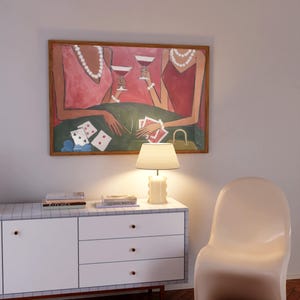 May include: An abstract painting depicts two figures in pink dresses playing cards and drinking cocktails. The artwork is framed in a wooden frame and hangs above a white cabinet with a lamp and books. A white chair is next to the cabinet.