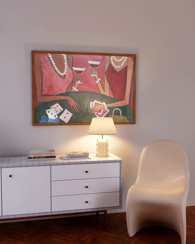 May include: An abstract painting depicts two figures in pink dresses playing cards and drinking cocktails. The artwork is framed in a wooden frame and hangs above a white cabinet with a lamp and books. A white chair is next to the cabinet.