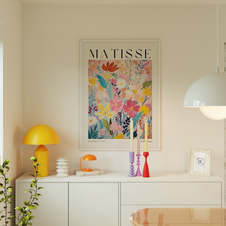 May include: A white framed print with the word "MATISSE" in black text and a colorful floral illustration. Three candle holders with white candles are on a white countertop with a yellow lamp, a stack of books, and a small framed picture.