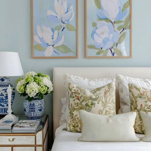 May include: Two framed floral art prints with light blue and white magnolia flowers hang above a bed with patterned pillows. A bedside table holds a lamp, books, and a vase of flowers, creating a serene bedroom scene.