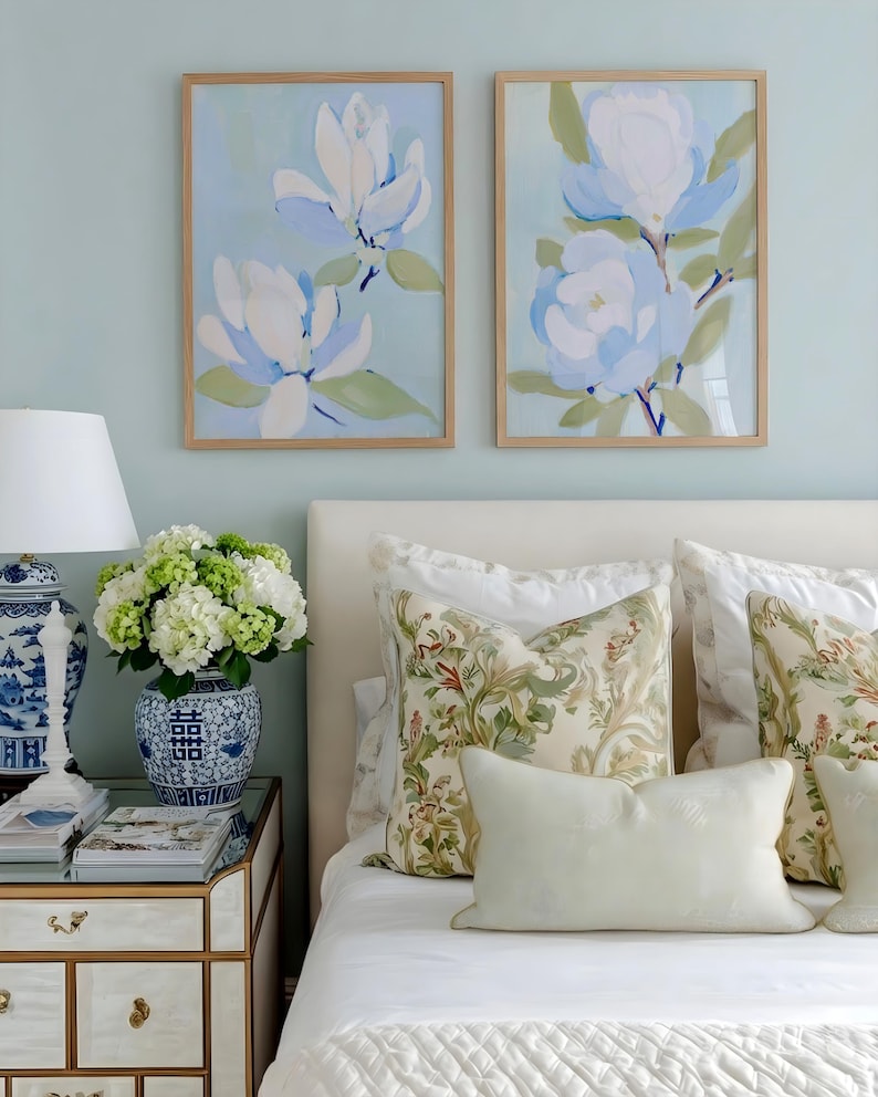 May include: Two framed floral art prints with light blue and white magnolia flowers hang above a bed with patterned pillows. A bedside table holds a lamp, books, and a vase of flowers, creating a serene bedroom scene.