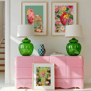 K&ouml;nnte beinhalten: Eine rosa Kommode mit zwei gr&uuml;nen Lampen und gerahmten Kunstdrucken. Die Kunst zeigt einen floralen L&ouml;wen, ein Herz mit Blumen und ein Herz mit V&ouml;geln. Eine blau gemusterte Vase steht oben auf der Kommode.