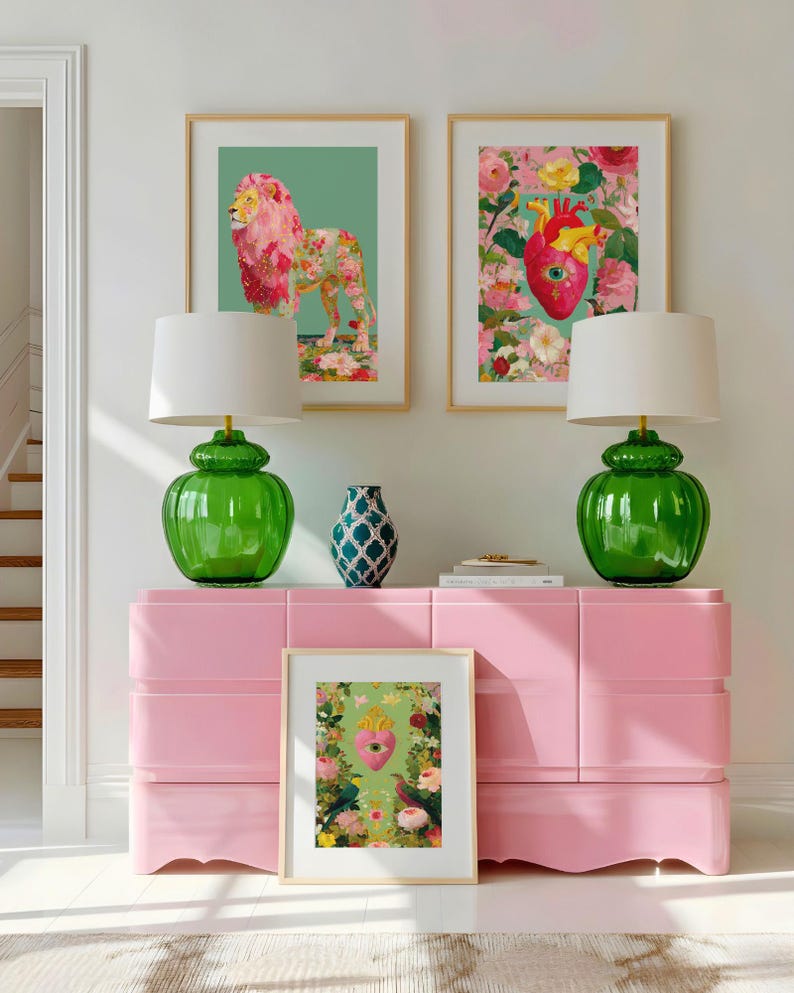K&ouml;nnte beinhalten: Eine rosa Kommode mit zwei gr&uuml;nen Lampen und gerahmten Kunstdrucken. Die Kunst zeigt einen floralen L&ouml;wen, ein Herz mit Blumen und ein Herz mit V&ouml;geln. Eine blau gemusterte Vase steht oben auf der Kommode.