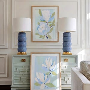 May include: Two blue ceramic lamps with white shades sit atop matching light green bedside tables. Two framed floral art pieces hang on the wall. The art features white and blue flowers with green leaves.