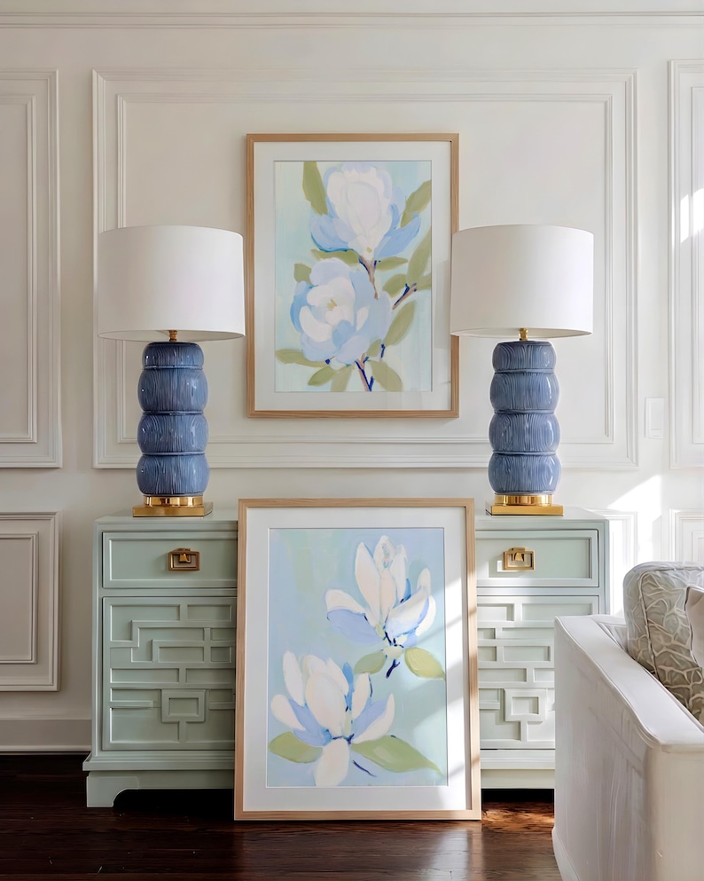 May include: Two blue ceramic lamps with white shades sit atop matching light green bedside tables. Two framed floral art pieces hang on the wall. The art features white and blue flowers with green leaves.