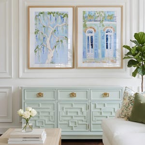 Puede incluir: Dos pinturas en acuarela enmarcadas de un &aacute;rbol y un edificio con contraventanas azules cuelgan sobre un aparador azul claro con herrajes dorados. Un sof&aacute; blanco y una mesa de centro de madera con un jarr&oacute;n de rosas blancas completan la decoraci&oacute;n de la habitaci&oacute;n.