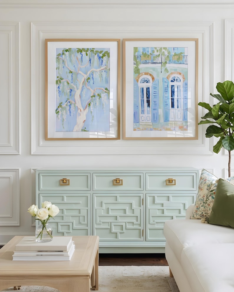 Puede incluir: Dos pinturas en acuarela enmarcadas de un &aacute;rbol y un edificio con contraventanas azules cuelgan sobre un aparador azul claro con herrajes dorados. Un sof&aacute; blanco y una mesa de centro de madera con un jarr&oacute;n de rosas blancas completan la decoraci&oacute;n de la habitaci&oacute;n.