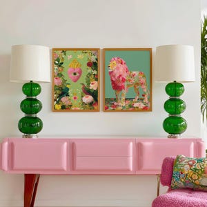 K&ouml;nnte beinhalten: Eine rosa Anrichte im Mid-Century-Modern-Design, flankiert von zwei gr&uuml;nen Glaslampen mit wei&szlig;en Schirmen. Dar&uuml;ber zwei gerahmte Kunstwerke, eines mit einem L&ouml;wen und das andere mit einem Herzen mit floralen Elementen. Rechts ein rosa Stuhl.