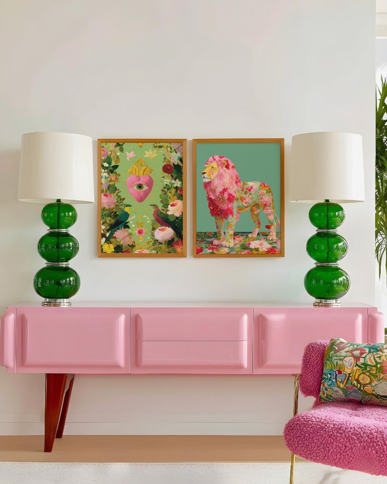 K&ouml;nnte beinhalten: Eine rosa Anrichte im Mid-Century-Modern-Design, flankiert von zwei gr&uuml;nen Glaslampen mit wei&szlig;en Schirmen. Dar&uuml;ber zwei gerahmte Kunstwerke, eines mit einem L&ouml;wen und das andere mit einem Herzen mit floralen Elementen. Rechts ein rosa Stuhl.