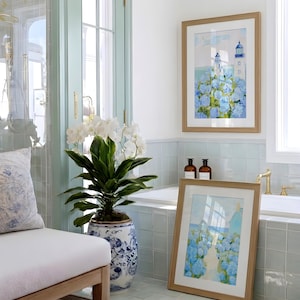 May include: A bathroom scene with a framed watercolor print of a lighthouse and blue flowers. A large potted plant with white flowers and a blue and white patterned pot is in the foreground. The room has a light blue and white color scheme.
