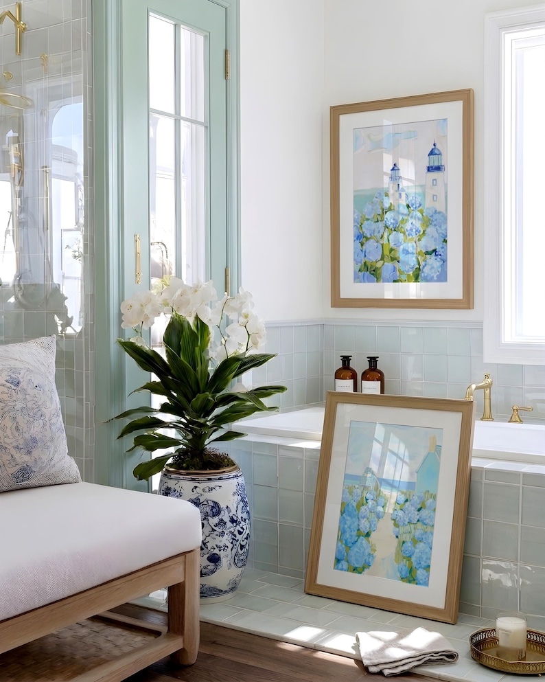 May include: A bathroom scene with a framed watercolor print of a lighthouse and blue flowers. A large potted plant with white flowers and a blue and white patterned pot is in the foreground. The room has a light blue and white color scheme.