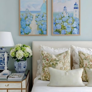 May include: Two framed landscape paintings with blue hydrangeas and a coastal scene. A white bed with floral pillows and a vase of white and green flowers on a mirrored nightstand. A blue and white lamp is also on the nightstand.