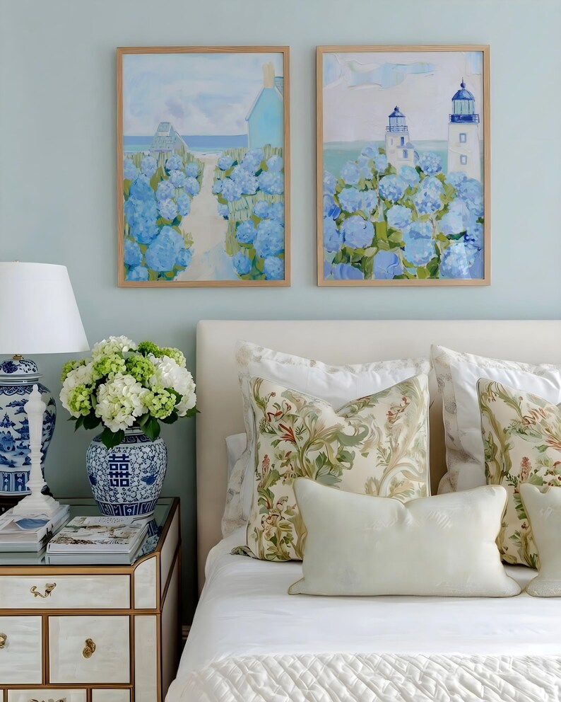 May include: Two framed landscape paintings with blue hydrangeas and a coastal scene. A white bed with floral pillows and a vase of white and green flowers on a mirrored nightstand. A blue and white lamp is also on the nightstand.