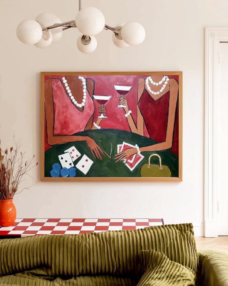 May include: An abstract painting in a wooden frame depicts two figures in red dresses, holding wine glasses. They are seated at a green table with playing cards, dice, and a purse. The artwork is set against a neutral wall, with a modern chandelier above.