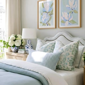 May include: A bedroom scene featuring a bed with light blue bedding, white sheets, and patterned pillows. Two framed floral art prints hang above the bed. A white lamp and a blue and white vase are on a nightstand.
