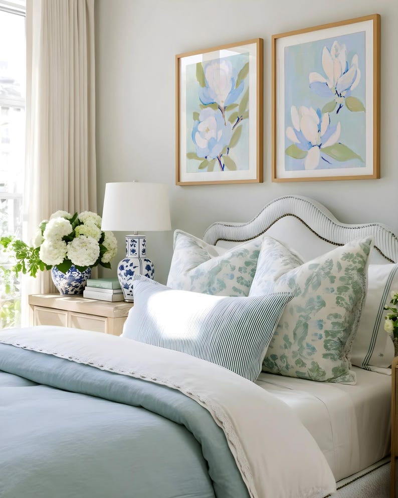 May include: A bedroom scene featuring a bed with light blue bedding, white sheets, and patterned pillows. Two framed floral art prints hang above the bed. A white lamp and a blue and white vase are on a nightstand.