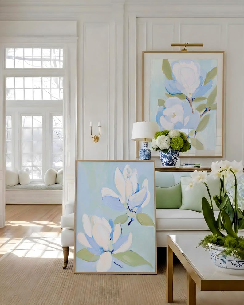 May include: Two framed floral paintings with light blue and white magnolia blossoms. The paintings are displayed in a bright, airy living room with a white sofa, green pillows, and a white coffee table with gold accents. The room has a neutral color palette.