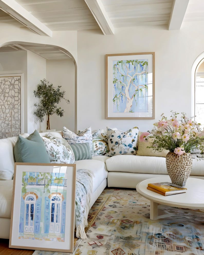 Puede incluir: Un sal&oacute;n luminoso y espacioso con un sof&aacute; esquinero blanco, cojines decorativos y una mesa de centro redonda. Dos pinturas de acuarela enmarcadas se apoyan en el sof&aacute;. Un jarr&oacute;n con flores rosas est&aacute; sobre la mesa.