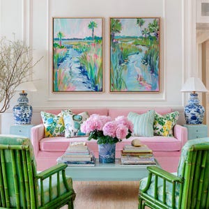 May include: Two rectangular landscape paintings in light wood frames featuring colorful marsh scenes with herons, palm trees, and water. The artwork displays vibrant brushstrokes in shades of blue, green, and pink, mounted on a white wall above a pink sofa.