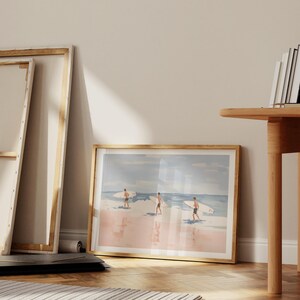 May include: A framed watercolor painting of three people surfing on a pink beach with blue water and a light blue sky.