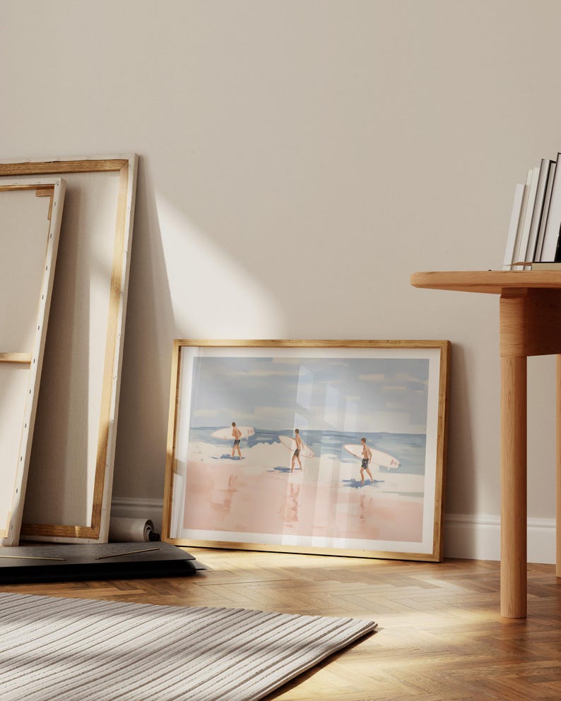 May include: A framed watercolor painting of three people surfing on a pink beach with blue water and a light blue sky.