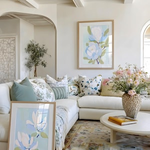 May include: A bright, airy living room with a white sectional sofa, decorative pillows, and a round coffee table. Two framed floral art prints in light wood frames lean against the sofa and hang on the wall. A vase of pink flowers sits on the table.