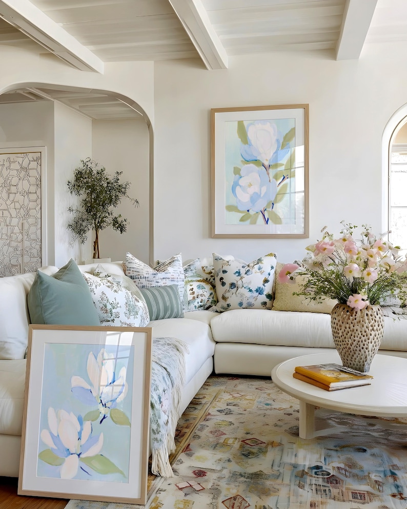 May include: A bright, airy living room with a white sectional sofa, decorative pillows, and a round coffee table. Two framed floral art prints in light wood frames lean against the sofa and hang on the wall. A vase of pink flowers sits on the table.